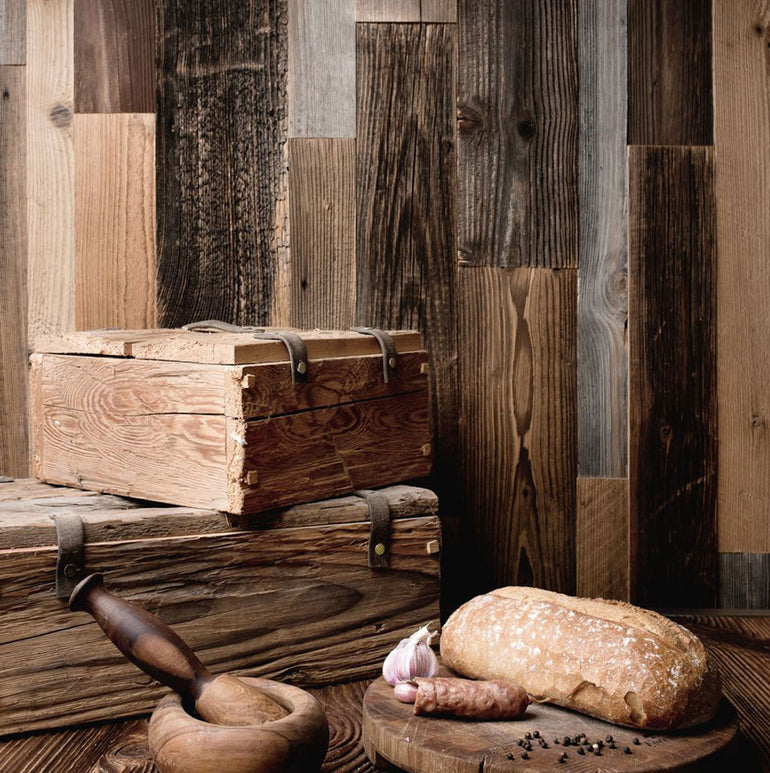 Une scène de cuisine rustique présente Parement Bois Antik de Wood & Wall Decor™ : deux boîtes en bois empilées, un mortier et un pilon, des saucisses, du pain, de l'ail et des grains de poivre posés sur une table avec un revêtement en planches de bois saisissant.