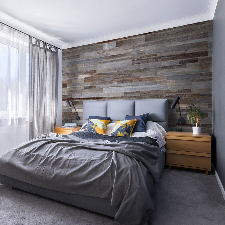 Une chambre à coucher moderne présente un mur d'accent Parement Bois Anthracite de Wood & Wall Decor™, un grand lit avec une literie grise et des oreillers colorés, deux tables de nuit en bois avec des lampes, une plante et des rideaux gris transparents pour un confort élégant.